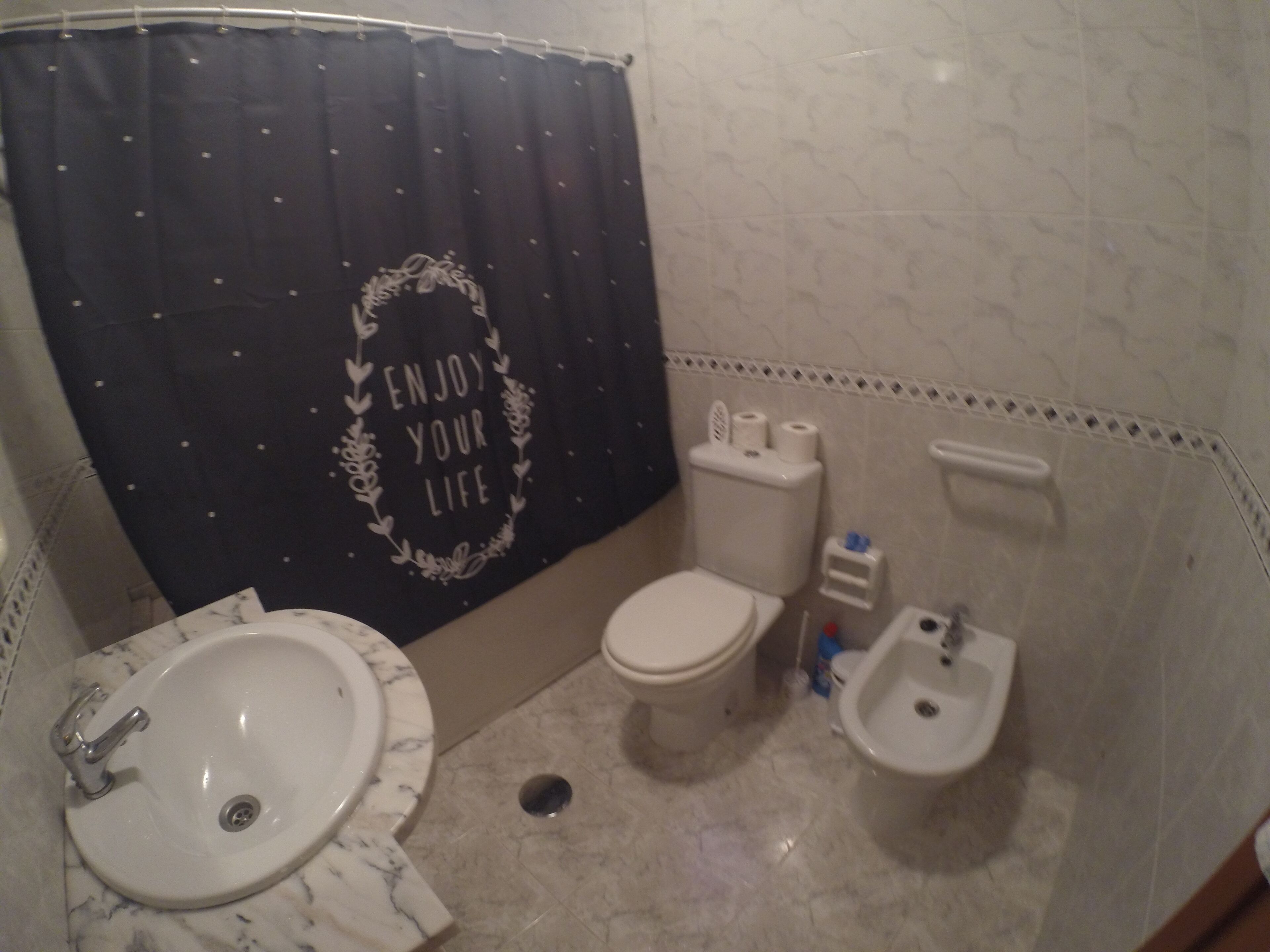 Une salle de bain avec un lavabo, des toilettes et un bidet blancs, un rideau de douche noir avec l’inscription « PROFITEZ DE LA VIE », et un sol carrelé.