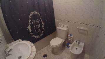 Une salle de bain avec un lavabo, des toilettes et un bidet blancs, un rideau de douche noir avec l’inscription « PROFITEZ DE LA VIE », et un sol carrelé.