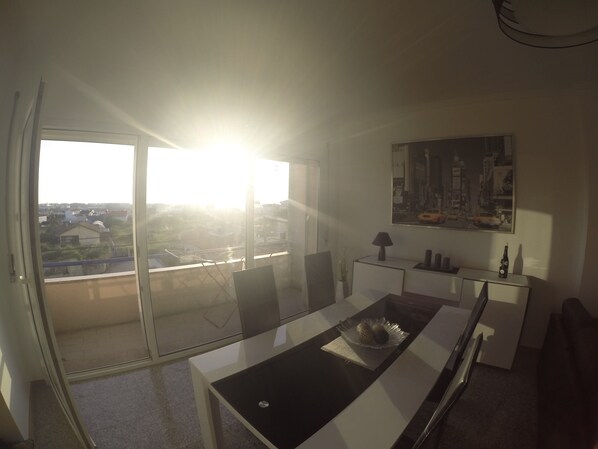 A modern kitchen with a glass door leading to a balcony, a dining table with a black top, and a cityscape view.