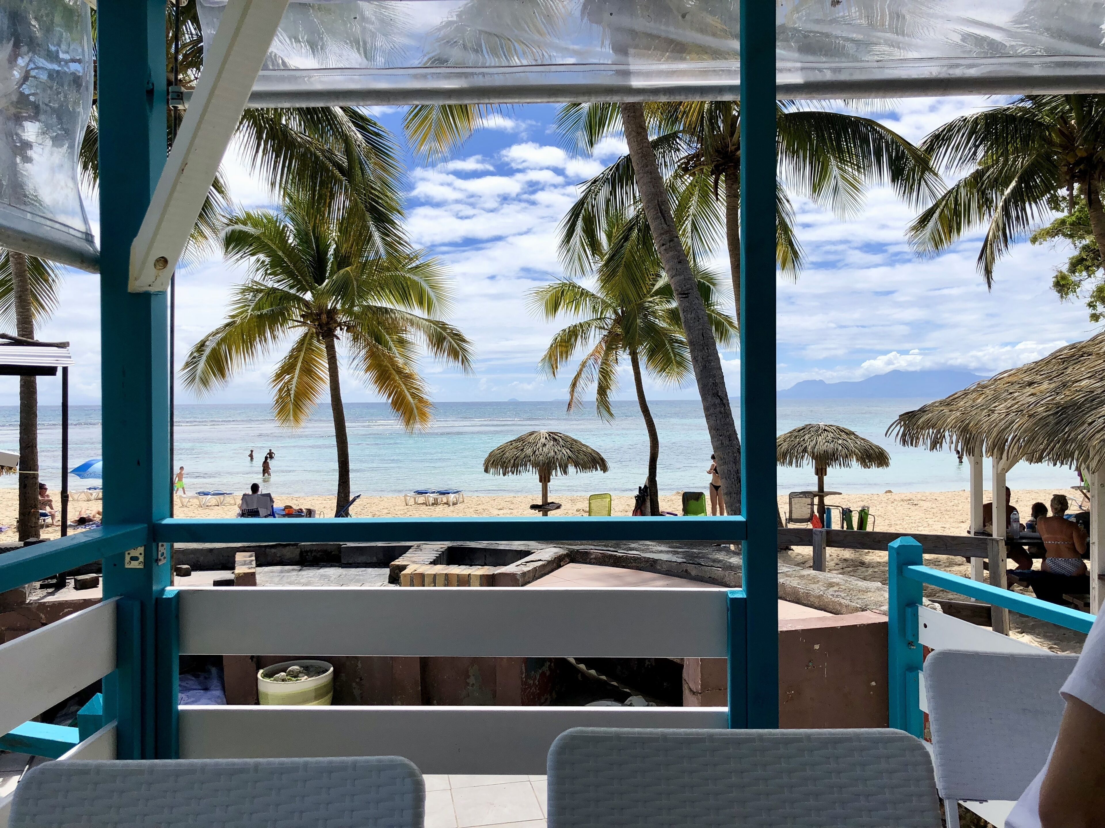 Vue sur la plage depuis un espace de repos abrité, agrémenté de palmiers et de parasols de plage.