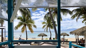 A beach view from a covered seating area with palm trees and beach umbrellas. - Panoramic sea view villa - private beach access (Saint-François)