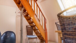Eine Treppe mit hölzernen Geländern, ein Korbstuhl und ein Metallstuhl in einem Zimmer mit hellem Boden und einer weißen Tür.