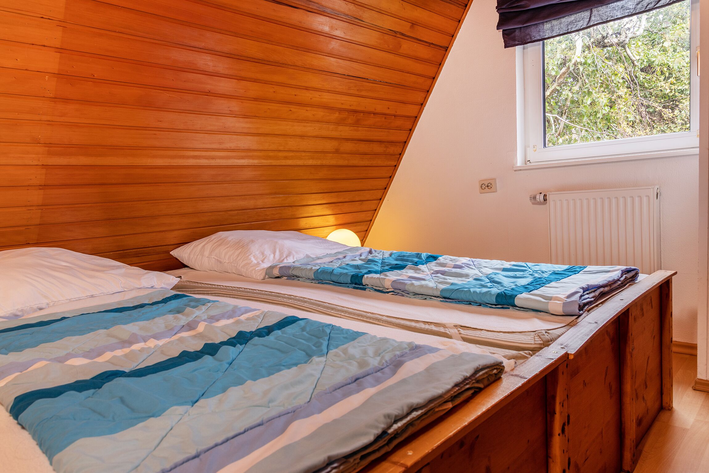 Slaapkamer met houten plafond, raam met uitzicht op bomen, radiator en bed met gestreept beddengoed.