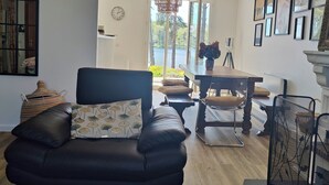A living room with a black leather sofa, a dining table, and a chandelier. There are framed pictures on the wall and a view of the outdoors through large windows.