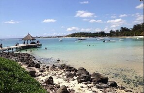 A beach with clear turquoise water, a thatched-roof structure, and boats in the distance.