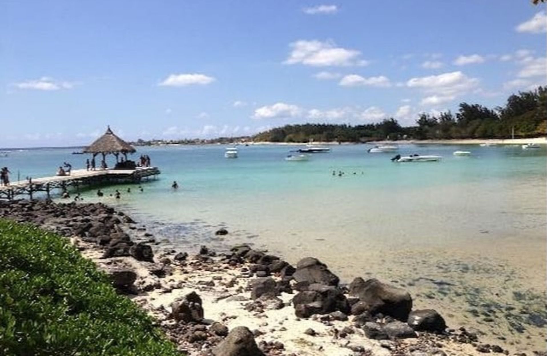 Une plage à l’eau turquoise et limpide, une structure à toit de chaume et des bateaux visibles au loin.