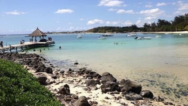 A beach with clear turquoise water, a thatched-roof structure, and boats in the distance.