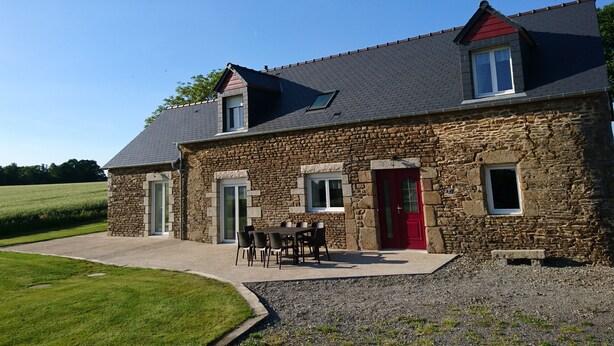 A stone house with a red door, a small balcony, and a patio with a table and chairs.