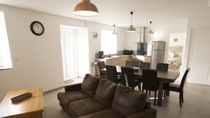 A modern living room with a brown leather sofa, a wooden coffee table, and a dining area with a dark wooden table and chairs. A kitchen with stainless steel appliances is visible in the background.