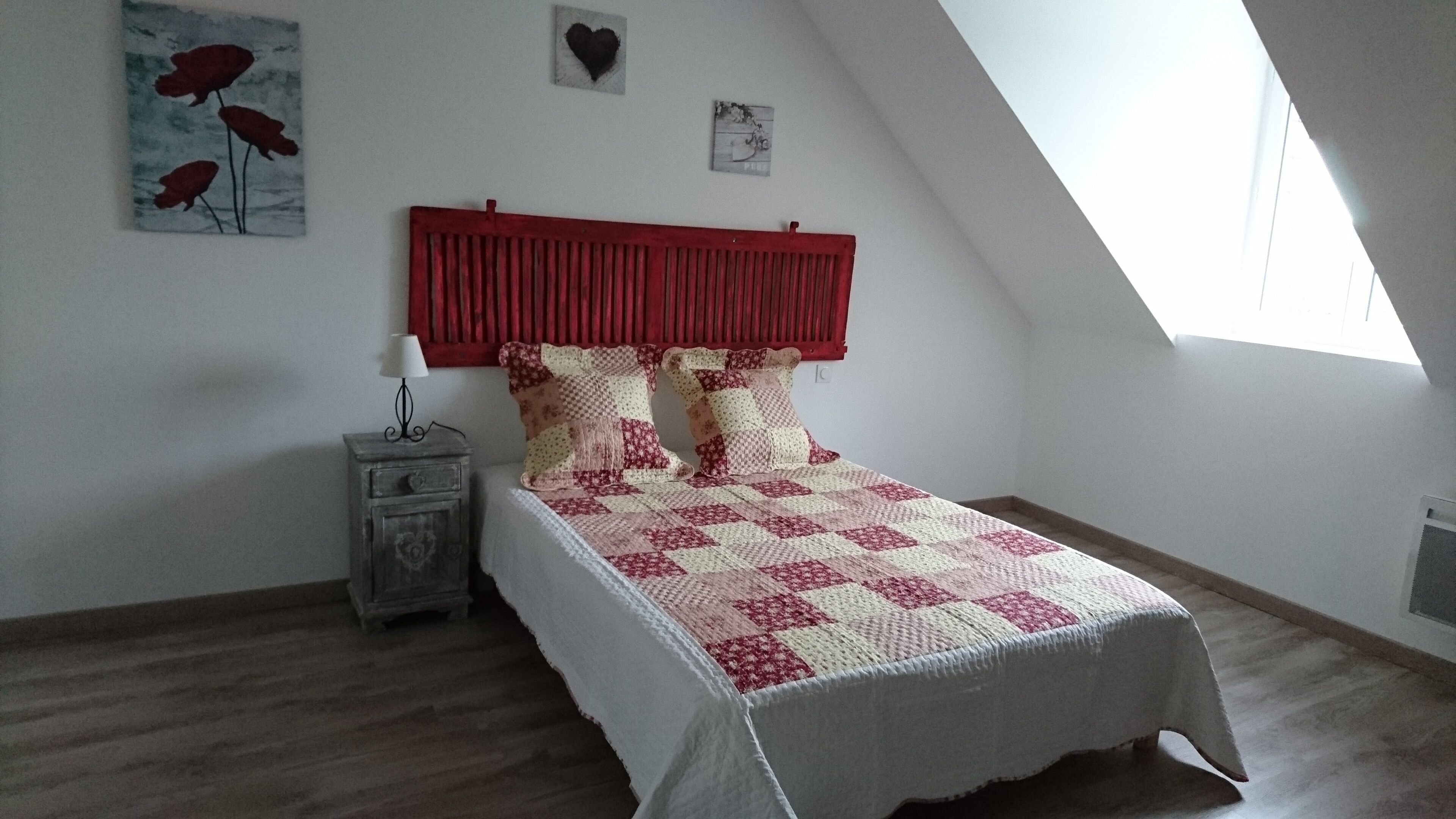 A bedroom with a sloped ceiling, a red headboard, a quilted bedspread, a nightstand with a lamp, and a small wall art piece.