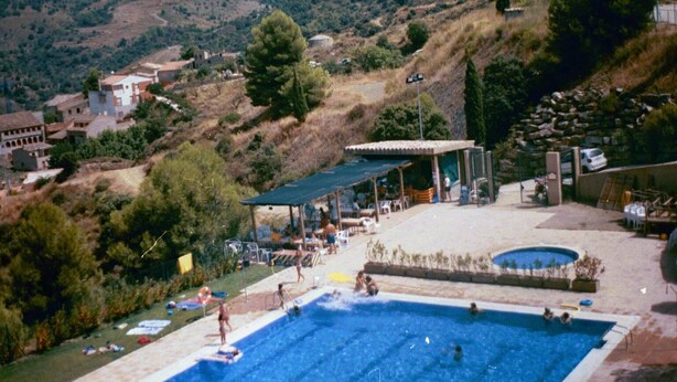 Una piscina con gente alrededor, una zona cubierta con mesas y sillas, y vistas a un paisaje montañoso con edificios al fondo.