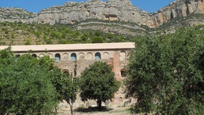 Un edificio histórico con ventanas de arco y tejado de tejas, rodeado de árboles y montañas rocosas.