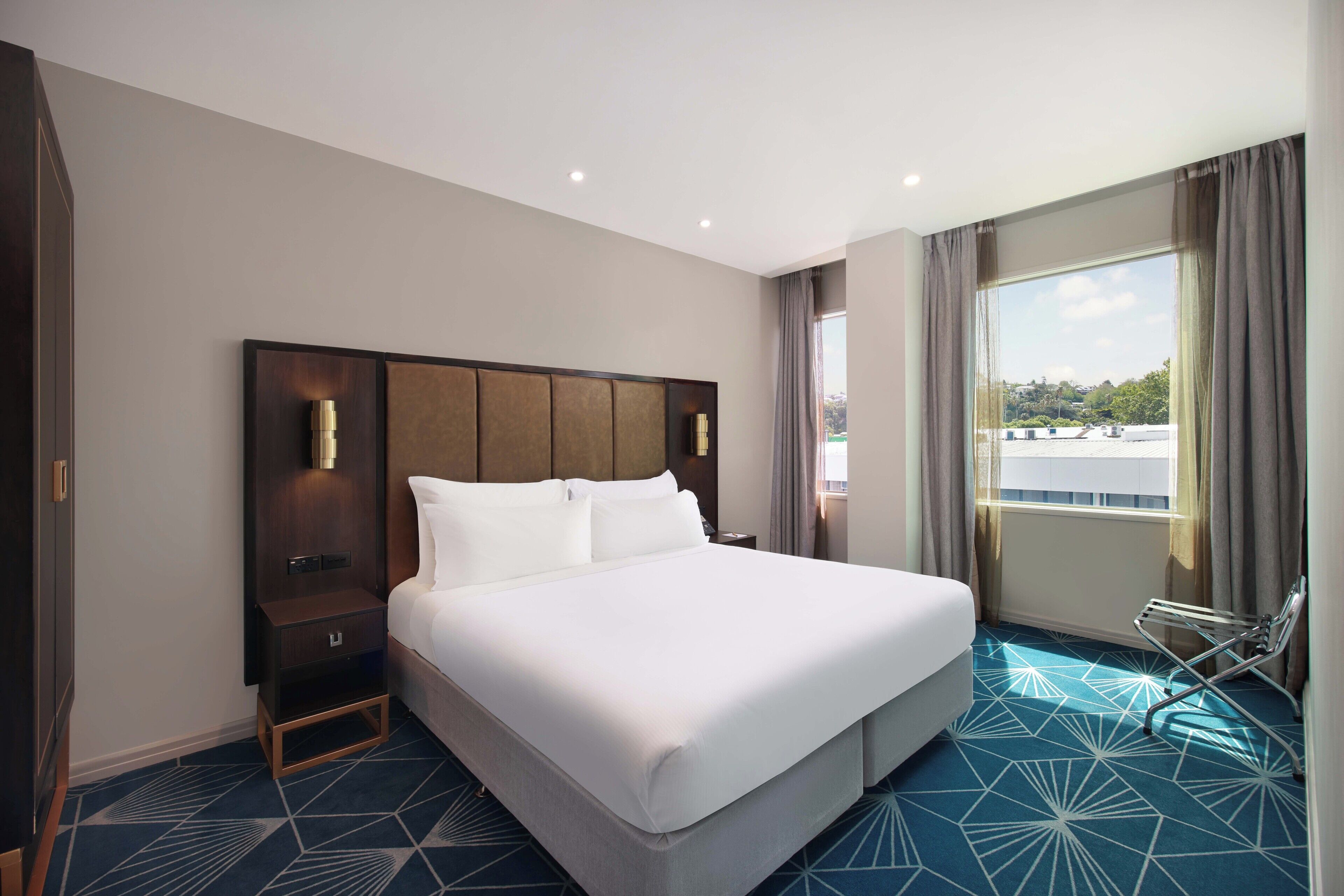 A hotel room with a large bed, a nightstand, a wardrobe, and a view of the city through a window.