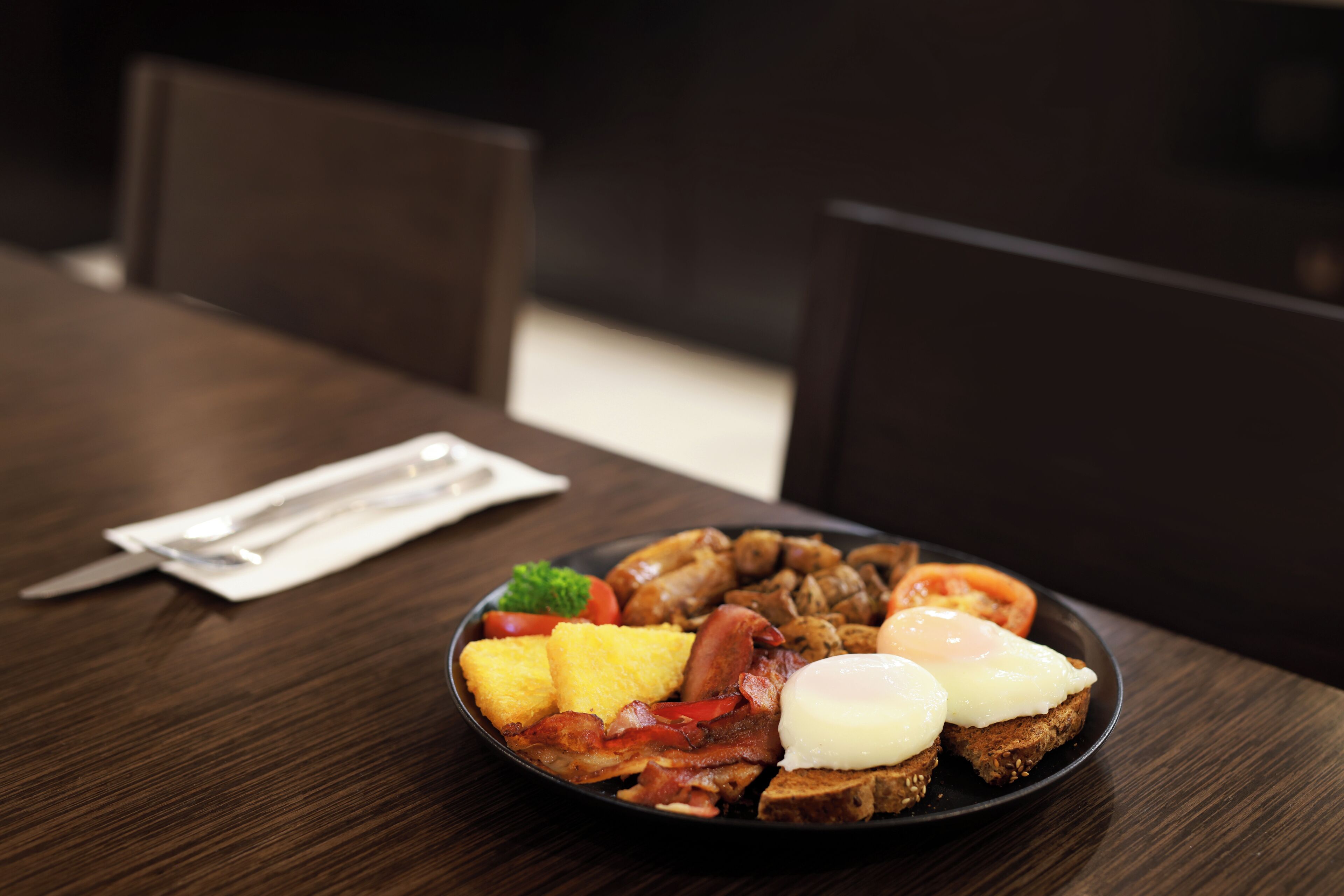 Une assiette de petit-déjeuner avec des œufs, du bacon, des tomates et des toasts, posée sur une table en bois foncé.