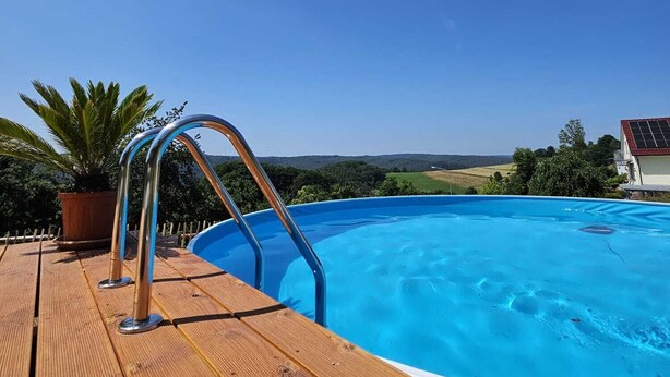 An outdoor swimming pool with a wooden deck and metal ladder, surrounded by greenery and a clear sky. - Apartment at Landhof Rothenberg - terrace with views of meadows and forests (Rothenberg)