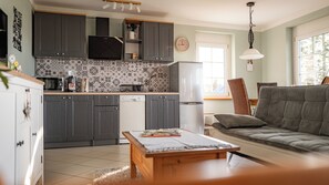 A modern kitchen with dark cabinets, a dishwasher, and a refrigerator. There is a dining area with a sofa and a table.