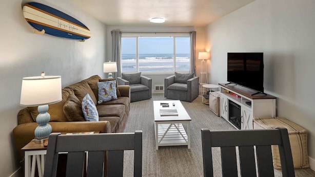 Sala de estar con sofá, dos sillones, una mesa de centro, televisor y chimenea. Amplias ventanas ofrecen vista al océano.
