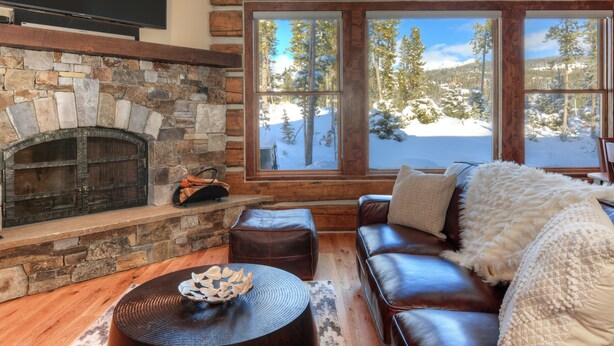 Ein gemütliches Wohnzimmer mit Kamin aus Stein, Ledersofa und großen Fenstern mit Blick auf eine verschneite Landschaft.