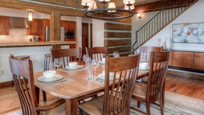 Ein Essbereich mit einem Holztisch, gedeckt für vier Personen, umgeben von Holzstühlen. Eine Chandelier hängt über dem Tisch, und im Hintergrund ist eine Treppe mit Geländer zu sehen.