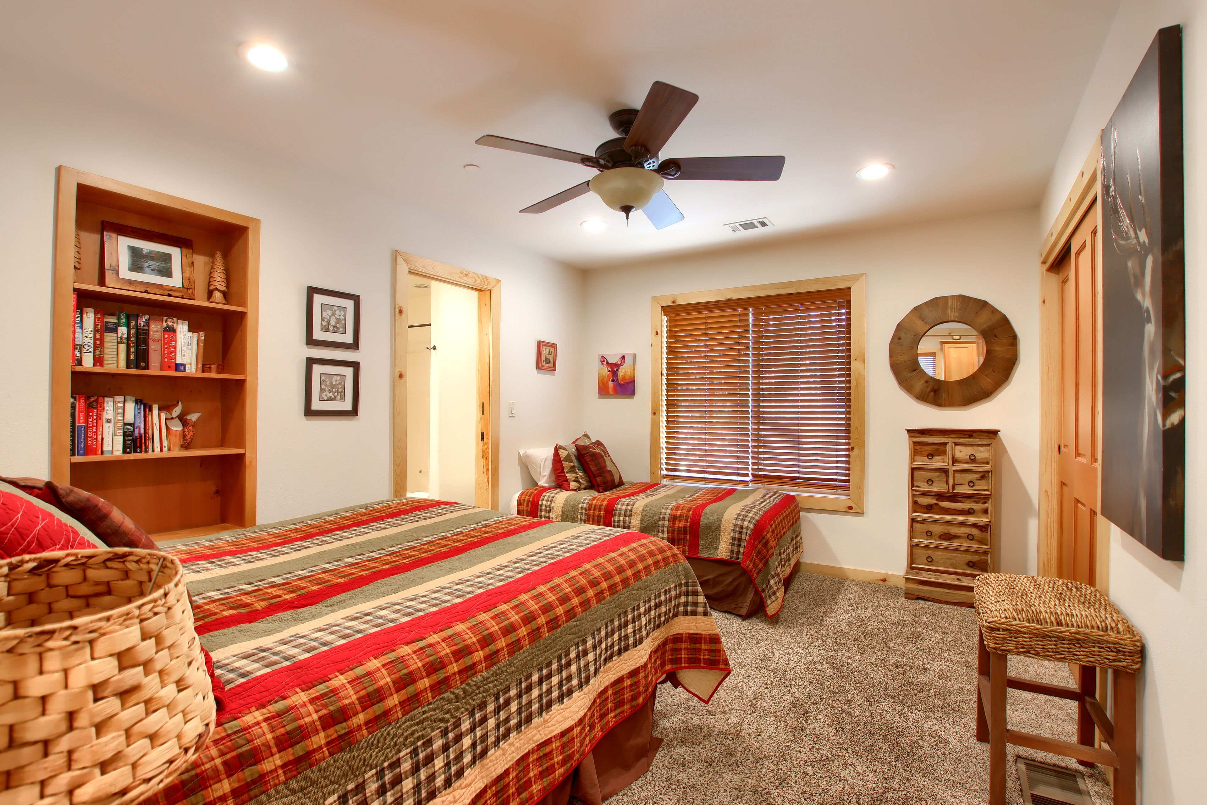 A bedroom with two beds, a ceiling fan, a bookshelf, and a window with blinds.