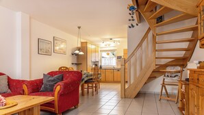 Ein gemütliches Wohnzimmer mit einem roten Sofa, einem Holztisch und einem Essbereich mit Tisch und Stühlen. Im Hintergrund ist eine Küche mit Holzschränken und eine Treppe zu sehen.