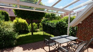 Eine überdachte Terrasse mit Tisch und Stühlen, umgeben von einer Ziegelmauer und Pflanzen.