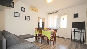 A living room with a gray sofa, a dining table with chairs, a TV on a stand, and a door leading to another room.