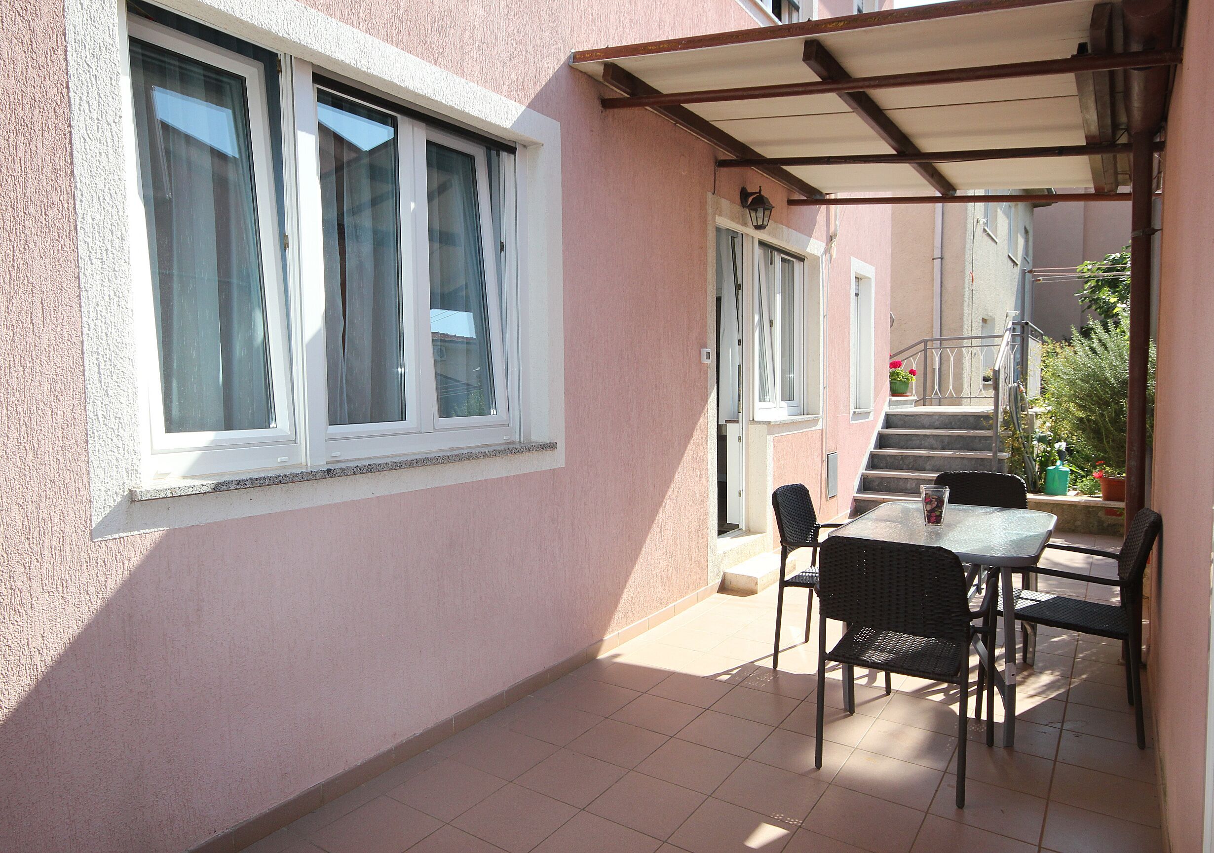 Une terrasse couverte avec une table et des chaises, une fenêtre avec des rideaux et un escalier menant à un étage supérieur.