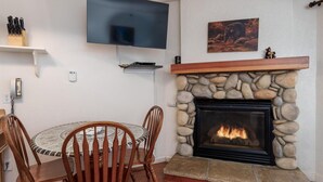 Ein gemütliches Wohnzimmer mit Kamin aus Stein, einem runden Esstisch mit Stühlen, einem wandmontierten Flachbildfernseher und einem Bärenbild an der Wand.