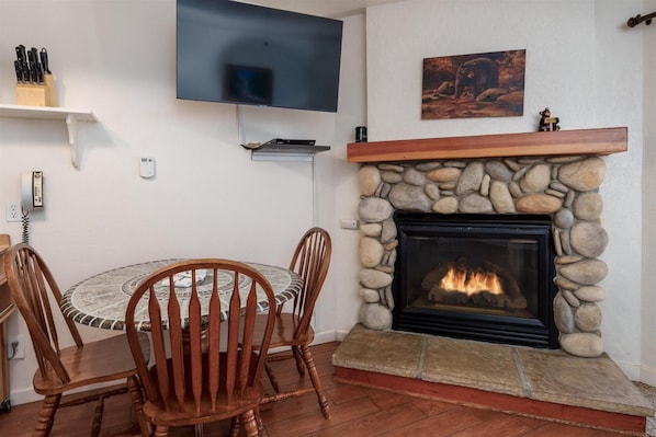 A cozy living room with a stone fireplace, a round dining table with chairs, a mounted flat-screen TV, and a bear-themed painting on the wall.