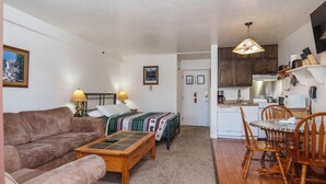 Ein gemütliches Wohnzimmer mit braunem Sofa, einem Holztisch und einem Bett mit einer grün-weißen Decke. Es gibt eine Essecke mit Holztisch und -stühlen sowie eine Küche mit Holzschränken und einem Herd.