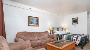 Ein Zimmer mit einem braunen Sofa, einem Holztisch, einem Bett mit grüner Steudecke und zwei Lampen.