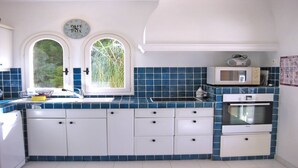 A kitchen with white cabinets, a blue tiled backsplash, a microwave, an oven, and two windows with greenery visible outside. - Villa with private pool - 10 people - Sea view - Air conditioning - WiFi - Sainte Maxime - Rated 4* (STE MAXIME)