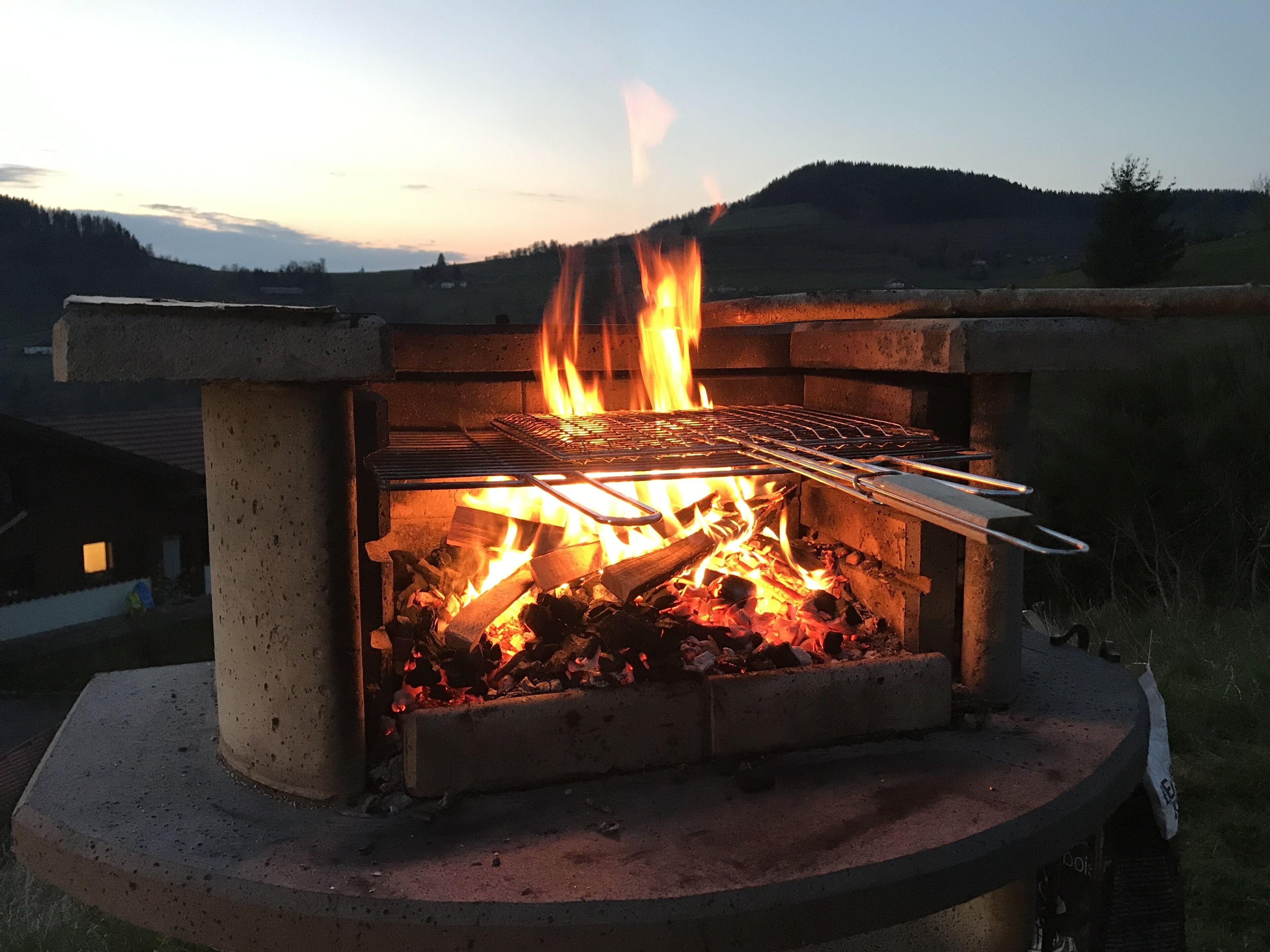 A wood-burning grill with flames, set in a concrete structure with a view of a hilly landscape in the background.