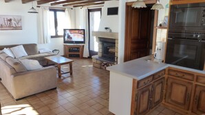 Ein Wohnzimmer mit Sofa, Couchtisch, Kamin, Fernseher und einer Küchenzeile mit Schränken und Geräten.