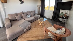 A living room with a sofa, a glass coffee table, a wicker chair, a TV, and a view of the outdoors.