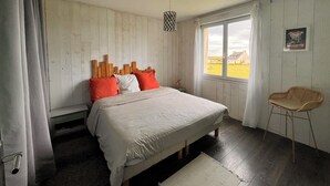 A bedroom with a wooden headboard, a bed with white bedding, a window with a view, a wicker chair, and a small table.