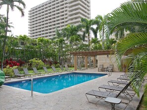 Piscina com espreguiçadeiras, rodeada por palmeiras e um edifício alto ao fundo.