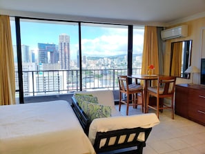 Quarto de hotel com uma cama grande, uma escrivaninha e uma cadeira. O quarto possui uma varanda com vista para a cidade.