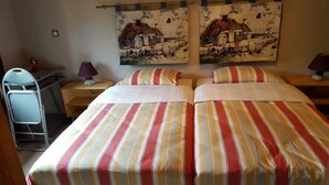 A hotel room with two beds, striped bedding, a desk with a lamp, and two wall art pieces depicting houses. - Atypical Cottage in the Chapel of the Miners (Sainte-Marie-Aux-Mines)