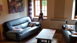 A living room with a black leather sofa, a coffee table, and a wooden floor. There is a window with a view of greenery outside. - Atypical Cottage in the Chapel of the Miners (Sainte-Marie-Aux-Mines)