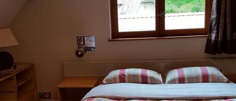 Een slaapkamer met een bed, een bureau, een stoel, een lamp, een raam met uitzicht op groen en een plafondlamp.