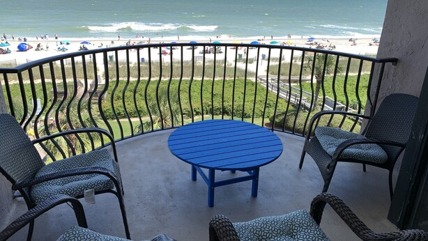 A balcony with wicker furniture and a blue table, overlooking a beach with umbrellas and people. - Amazing Views - Ocean Front 3BR Condo! (Myrtle Beach)