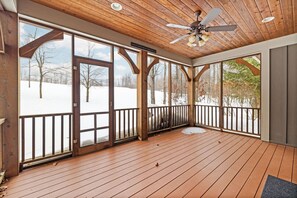 A covered wooden deck with a ceiling fan, large windows, and a view of a snowy landscape.