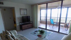 A living room with a sofa, a coffee table with a plant, a TV on a stand, and a sliding glass door leading to an outdoor balcony with chairs. - Gateway to Sanibel/Captiva 2/2 Ft. Myers Condo (Ft Myers)