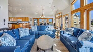 A spacious living room with a large blue sofa set, a round coffee table, and a view of the outside through large windows. - Oceanview Dog Friendly Home with Pool, Hot Tub, Elevator, Game and Media Rooms (Avon)
