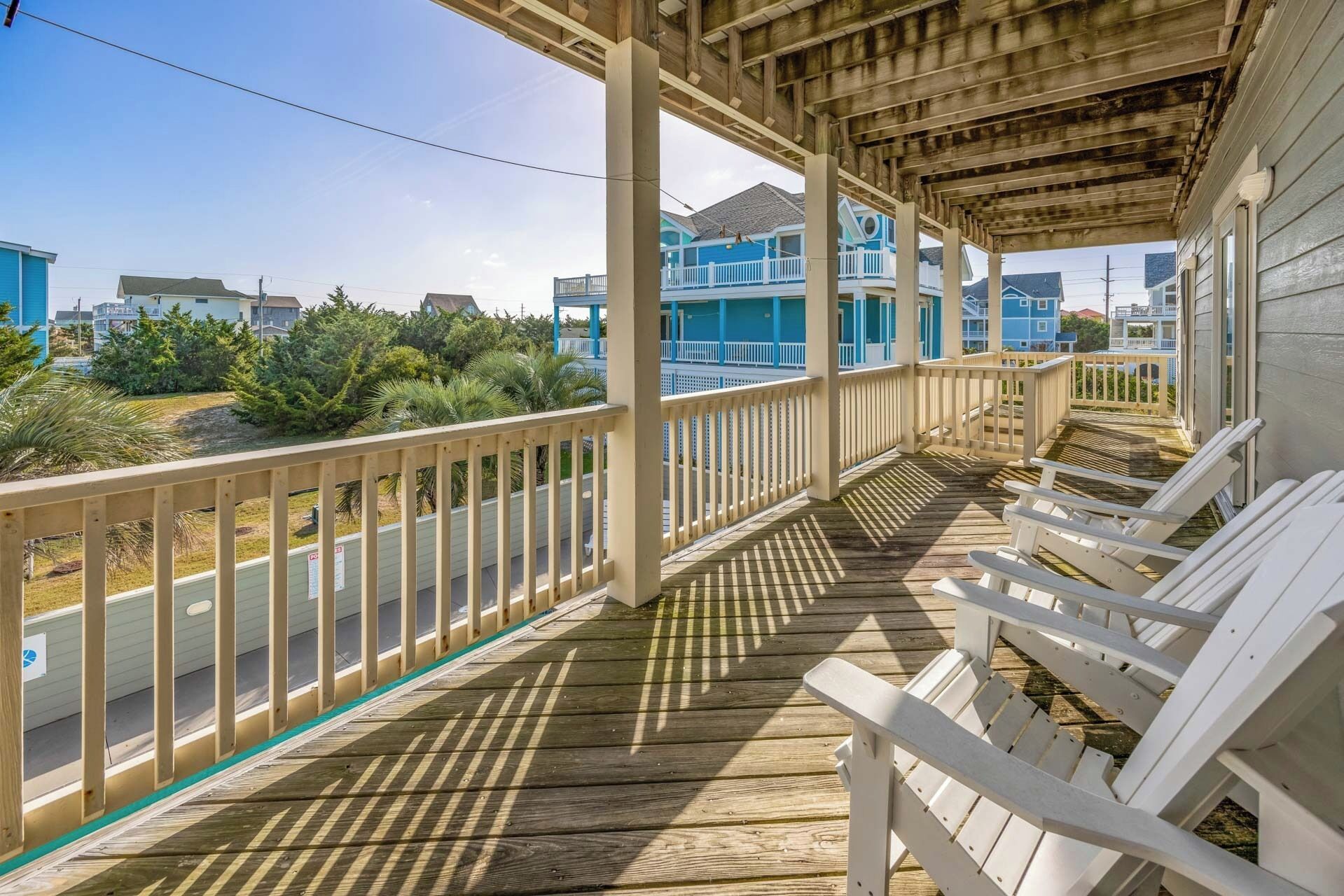 A wooden deck with white chairs, a covered area with a railing, and a view of blue buildings in the background.