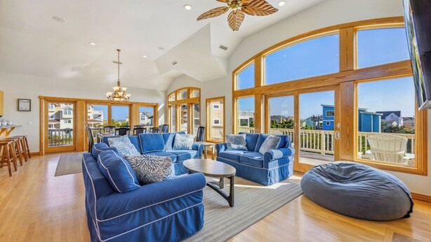 A spacious living room with blue sofas, a coffee table, and a bean bag. Large windows offer a view of the outside. - Oceanview Dog Friendly Home with Pool, Hot Tub, Elevator, Game and Media Rooms (Avon)