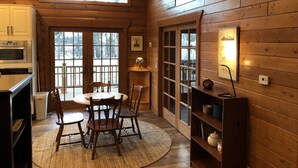 A cozy wooden cabin interior with a dining area, wooden walls, a large window, and a kitchen with a built-in oven.