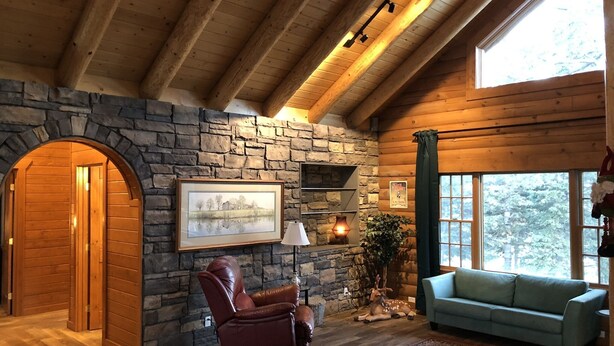 A log cabin interior with a stone fireplace, a reclining chair, a sofa, and a large window offering a view of the outdoors.